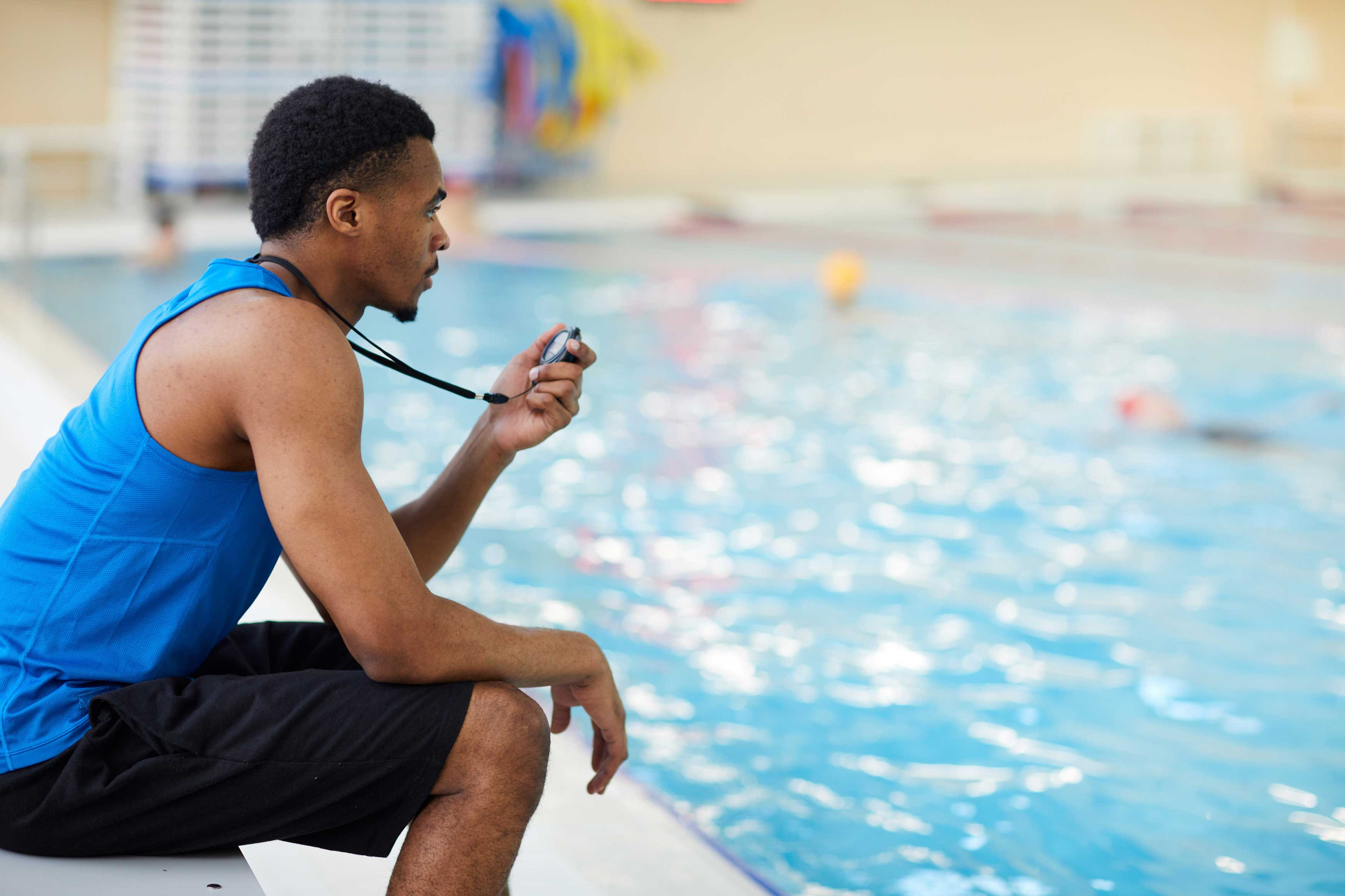 junger Mann sitzt am Rand eines Schwimmbeckens mit Stoppuhr in der Hand